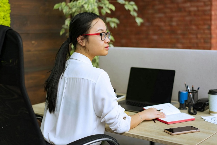 A woman in glasses sitting at a desk with a laptop and notebook, focused on applying 10 graphic design hacks to 10X social media growth.